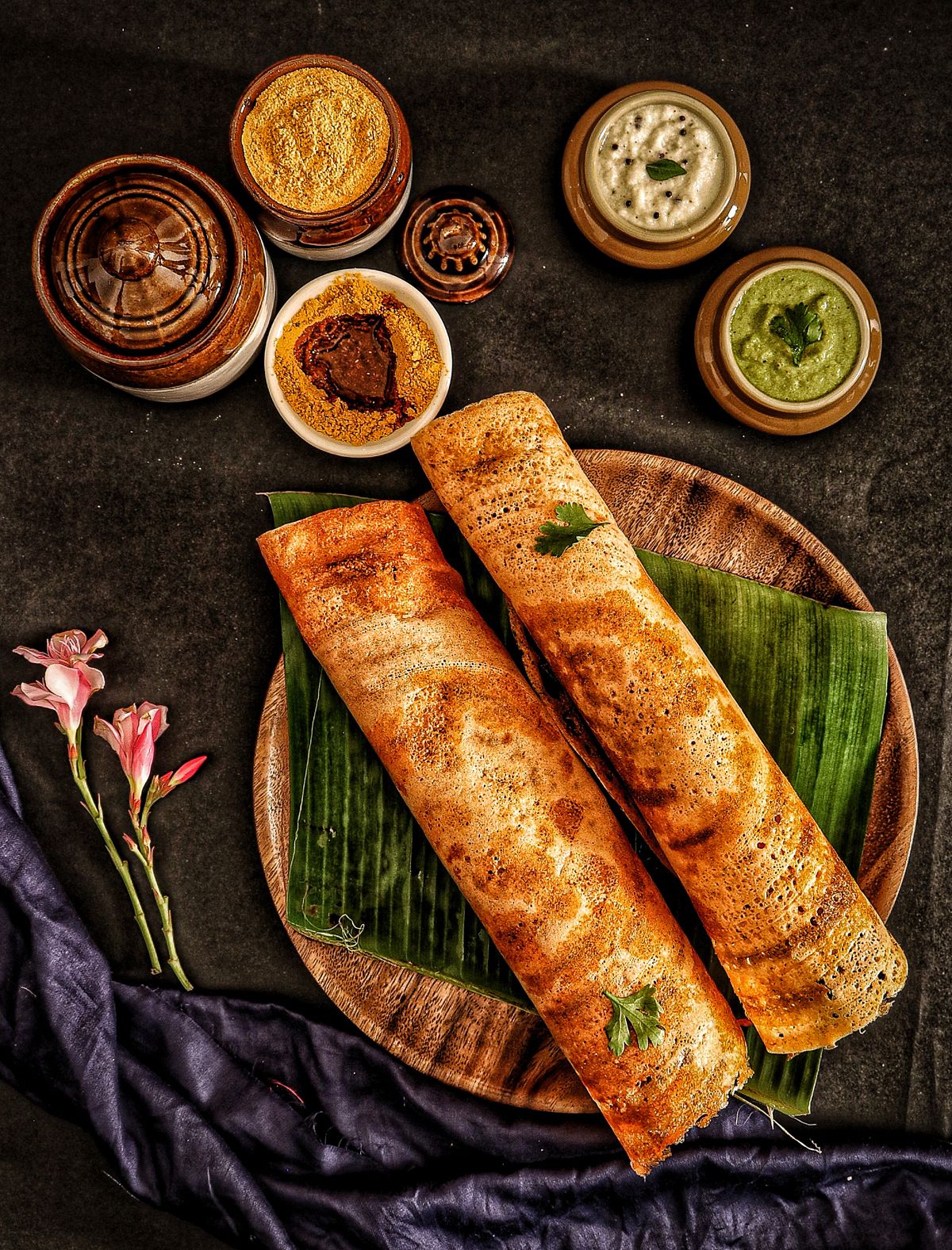 Two masala dosas on a banana leaf with chutneys and podi.